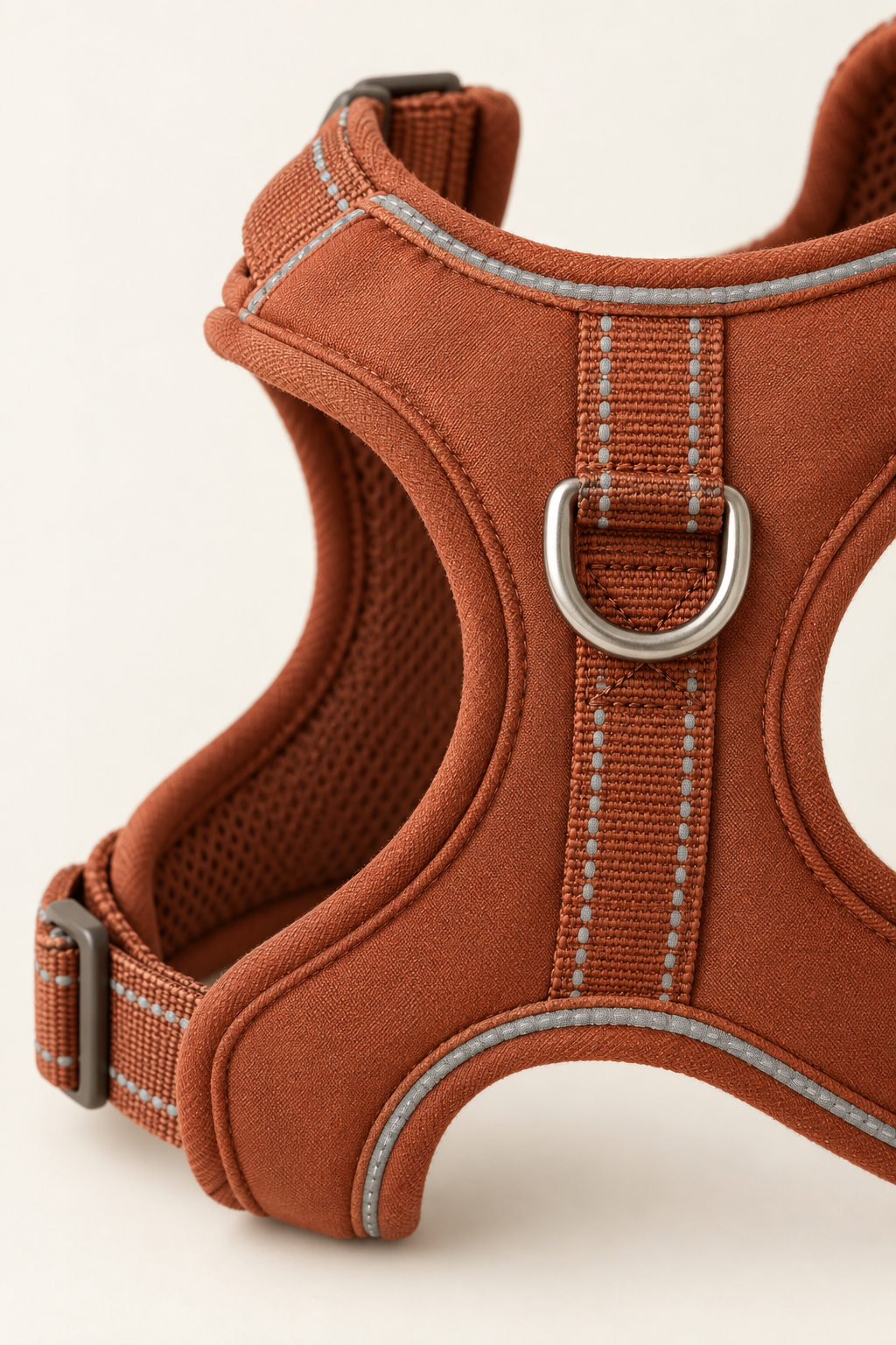 A close detail crop of the reflective stitching and metal D-ring on a terracotta dog harness on a warm cream background.