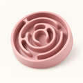 Round blush pink plastic slow feeder dog bowl with a raised concentric spiral maze moulded into the inside, photographed at a three-quarter angle