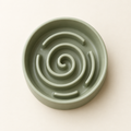 A pale sage green slow feeder pet bowl photographed from above showing the raised spiral maze in the base on a warm cream background.