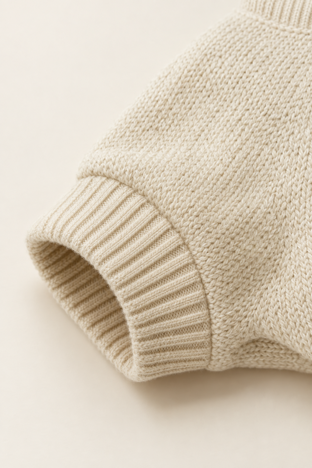 A close detail crop of the ribbed cuff and knit texture of a cream pet sweater on a warm cream background.